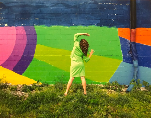 A female wearing green with her back to the viewer poses in front of a multi-colored wall