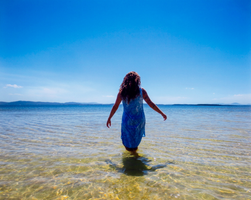 A female figure in a blue and white printed dress stands in a lake before a bright blue sky