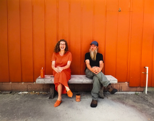 A smiling female figure wearing orange sits next to a bearded man on a wooden bench in front of an orange wall
