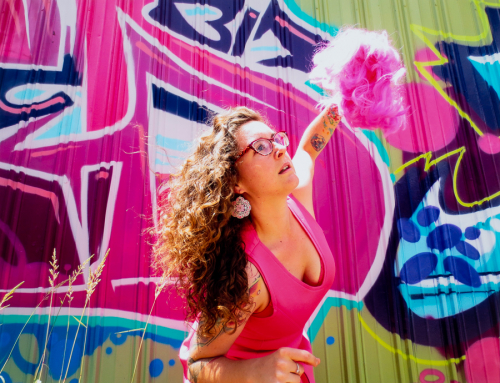 A female figure dressed in bright pink holding a pink pompom poses in front of a multi-colored wall of graffiti