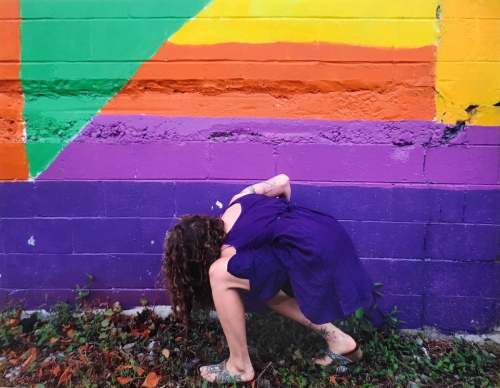 A female figure wearing purple crouches beside a multi-colored wall, the bottom of which is primarily purple