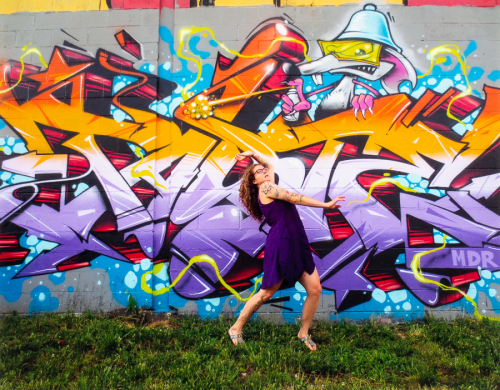 A female figure wearing purple poses in front of a multi-colored graffiti wall