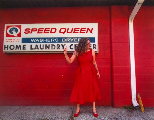 An image of a female in a red dress standing in front of a red wall and a sign for Speed Queen washers and dryers