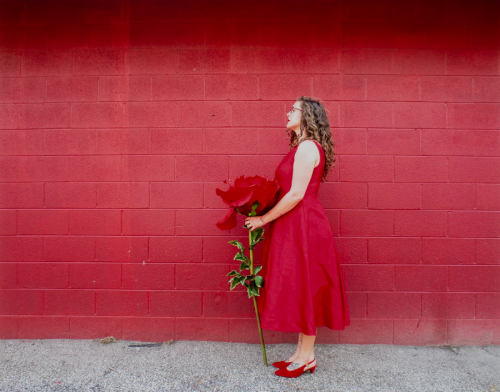A female figure in a red dress hold an oversized red flower in front of a red cinder block wall