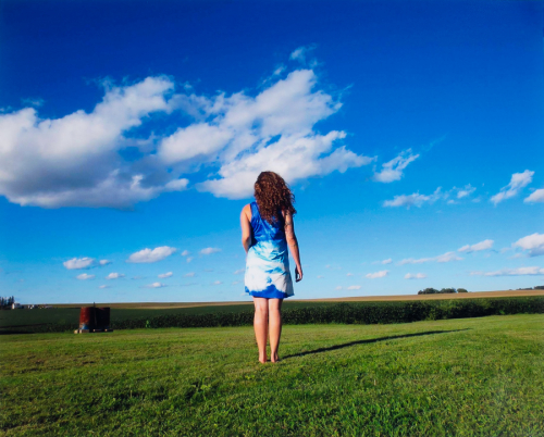 A female figure in a blue and white printed dress stands in a green field before a bright blue sky.