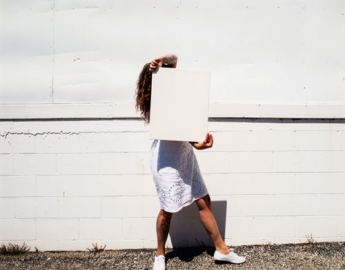 A figure in a white skirt covering her face with a white rectangular board stands in front of a white wall