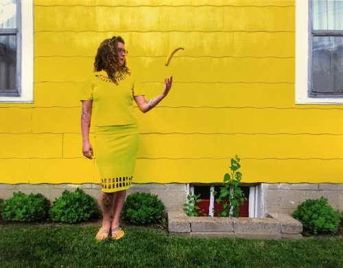 A female figure wearing yellow and tossing a banana in the air stands in front of the yellow wall of a house