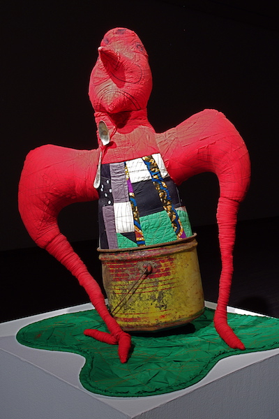 A red human-like sculpture in a rusted bucket an a green curvilinear pad.