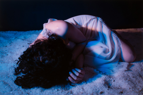 A female figure with long dark hair and in wash of blue light, lies curled up on a bed of large-crystal rock salt