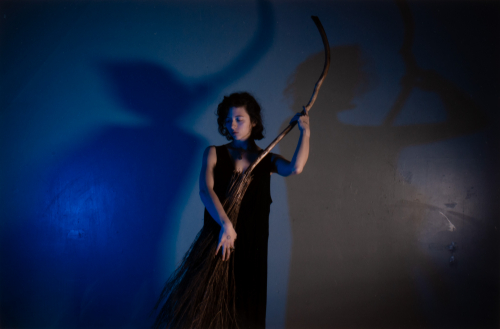 An image of a female figure holding a wooden broom standing against a wall in a wash of blue light