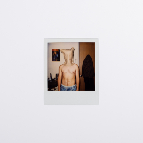 A self-portrait Polaroid photograph showing the shirtless artist in an interior space with a bag on his head