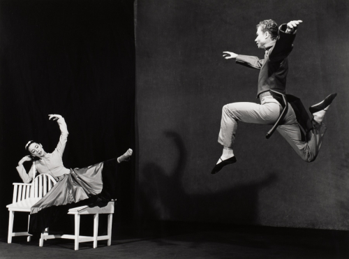 Woman and man dancing on dark stage. She is seated on a white bench with left arm and leg extended he is jumping.