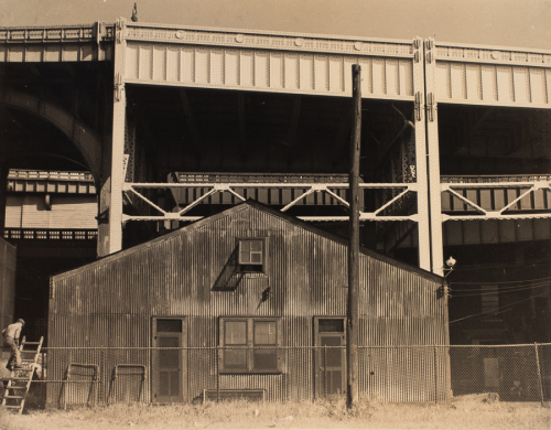 An image featuring a small building standing in front of a much larger steel structure. A male figure ascends a ladder 