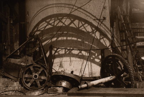 broken gears and other mechanical parts scattered on the floor in front of a wall with geometrical shadows from the girder above