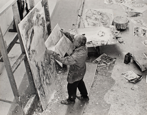View of man from steep angle above, in center of image, standing and holding a newspaper at arms length toward a canvas 