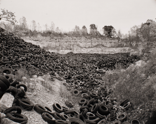 gigantic pile of rubber tires, grassy area along right side, higher cliff area with trees on top in background