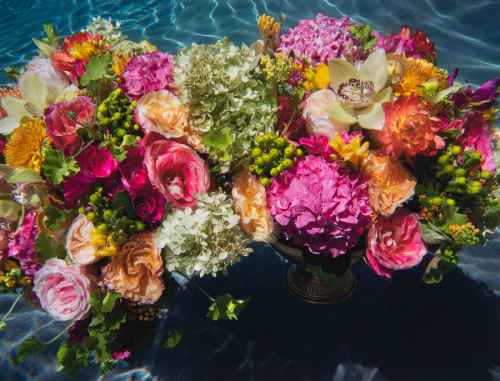 A large and colorful bouquet of flowers (roses, zinnias, hydrangeas, and a single orchid) in a metal vase as viewed underwater.