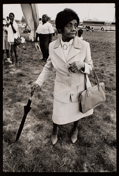 an elderly African American woman carrying an umbrella and purse standing outside with buses in the distance