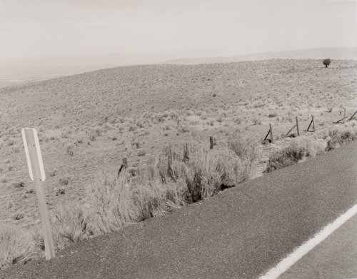 Roadway running diagonally across bottom; sign in lower left; fence posts along road and dry vegetation and hills in background