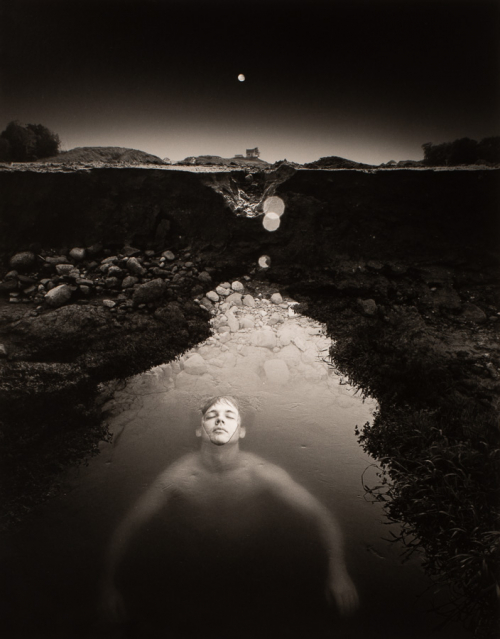 Black and white image of a moonlit landscape with a triangular pool of water and a partially submerged male figure.