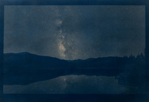 A cyanotype depicting a reflective lake in a mountainous landscape in front of a starry sky. 