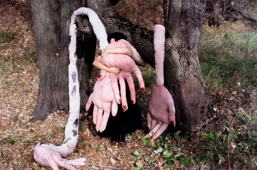 4 large soft-sculpture human hands, 2 of which are being hugged close to the body of a black-dressed female figure in the center