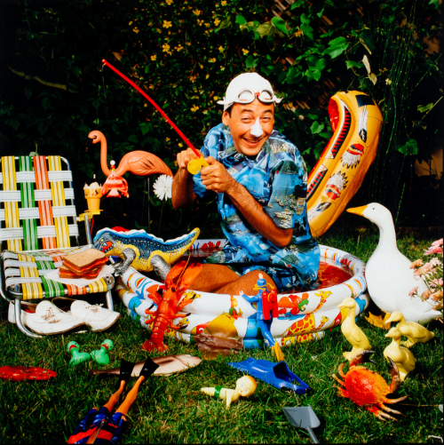 a man sitting in a child's swimming pool holding a child's fishing rod surrounded by numerous plastic toys and lawn chair