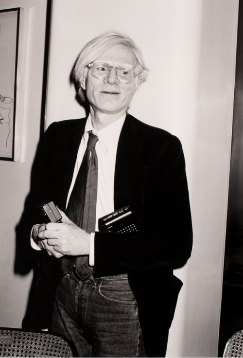  the artist wearing a suitcoat, tie, and eyeglasses and holding two black objects, probably a tape recorder and camera flash 