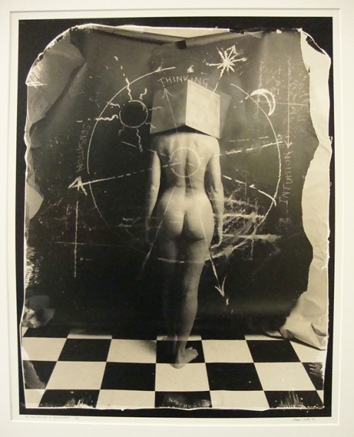A female nude wearing a cube on her head, standing on a black and white checkered floor
