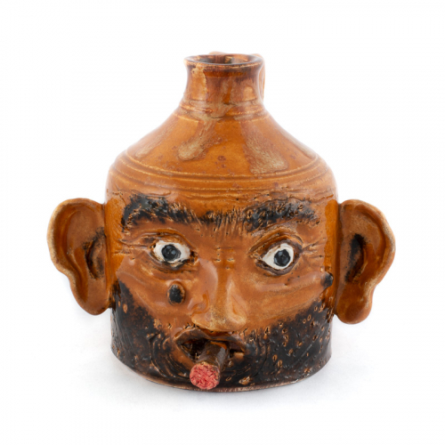  small face jug with handle, large ears, and lit cigar. glazed in a light brown with dark brown hair and a mole left of nose