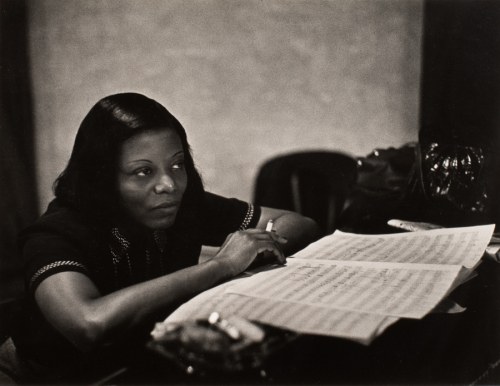 A woman seated and leaning on her elbows holding a cigarette at a bar table sheets of music are in front of her