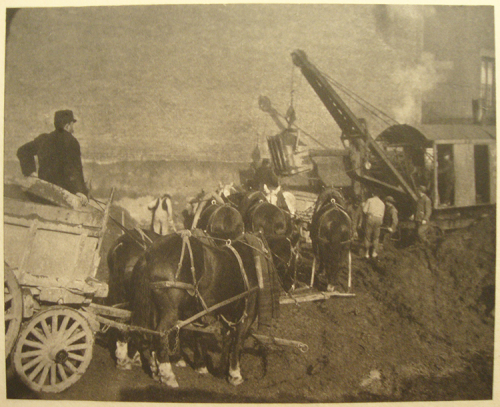 A black and white image of a horse-drawn carriage with an excavator in the distance