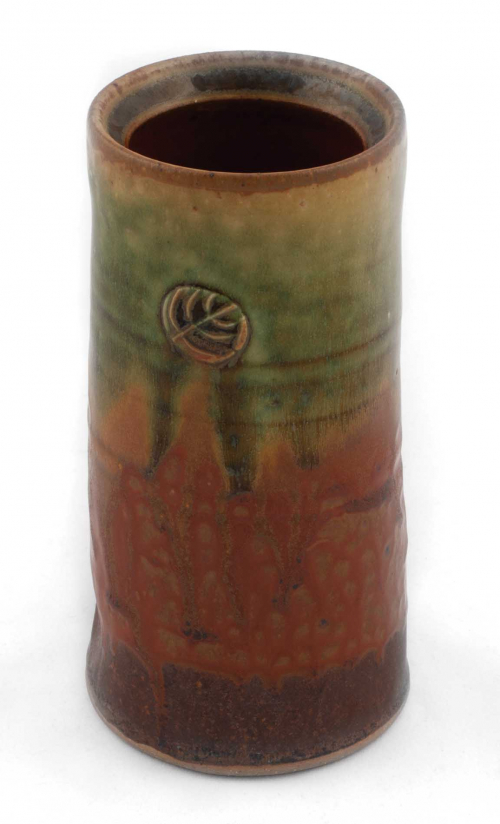 short cylindrical vase with a diagonal ridge in the mouth. Glazed in orange with green dripping from the rim and a raised leaf
