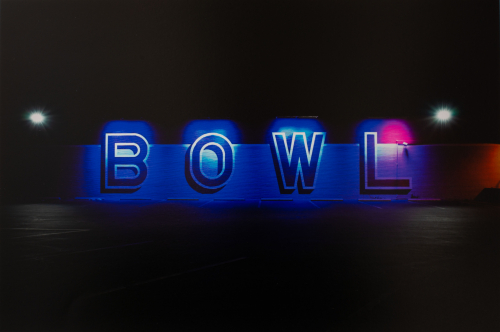 A photograph of the side of a bowling alley at night with blue and red lights illuminating a painted wall reading "BOWL".