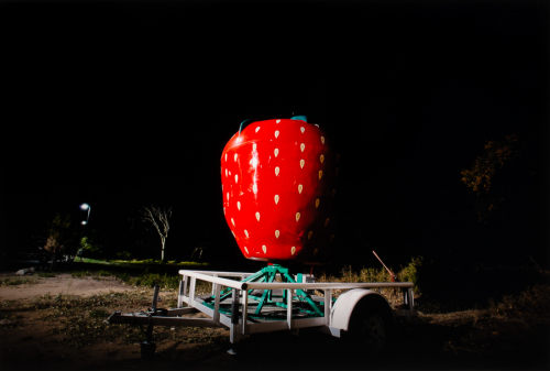  image shot outside at night featuring a large red strawberry sculpture on a green metal stand in a white trailer