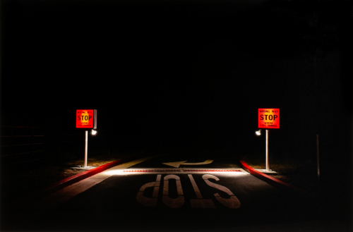 symmetrical composition of a road at night with the word "Stop" in the foreground and two brightly lit red-orange signs on sides