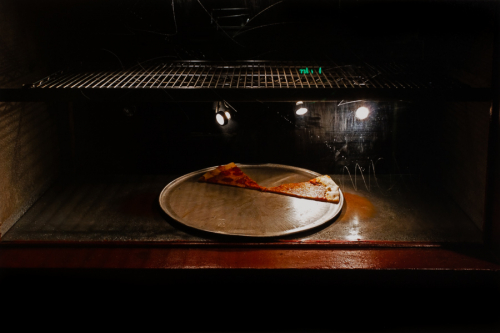 inside of a dark oven through a scratchy window, upper portion is a wire shelf and the lower is a pan with two slices of pizza
