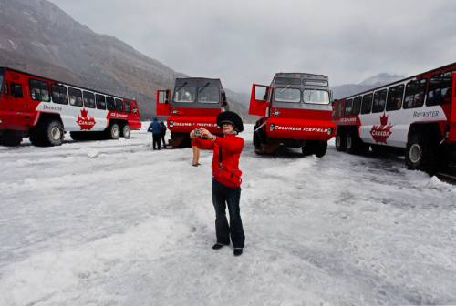 female wearing a red jacket and bag and a black helmet holds a camera in front of 4 large tourist vans on a field of ice.