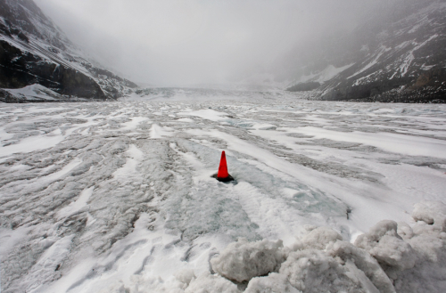 image of an icy passage between stony hills on an overcast day. The focal point is an orange traffic cone placed at center