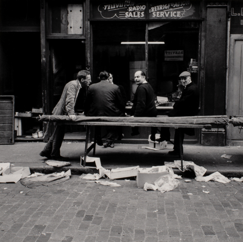 Five men in suits sit together on a littered curb in front of ruined storefronts.