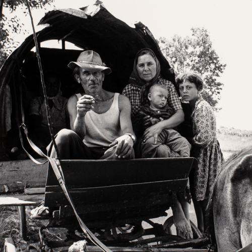 man, woman, and two children at the front of a wooden wagon, haunch of a horse in foreground and a kerchiefed woman in back