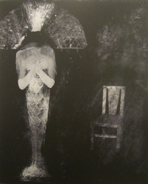 A black and white image with a mummy-like figure to the left and a chair to the right